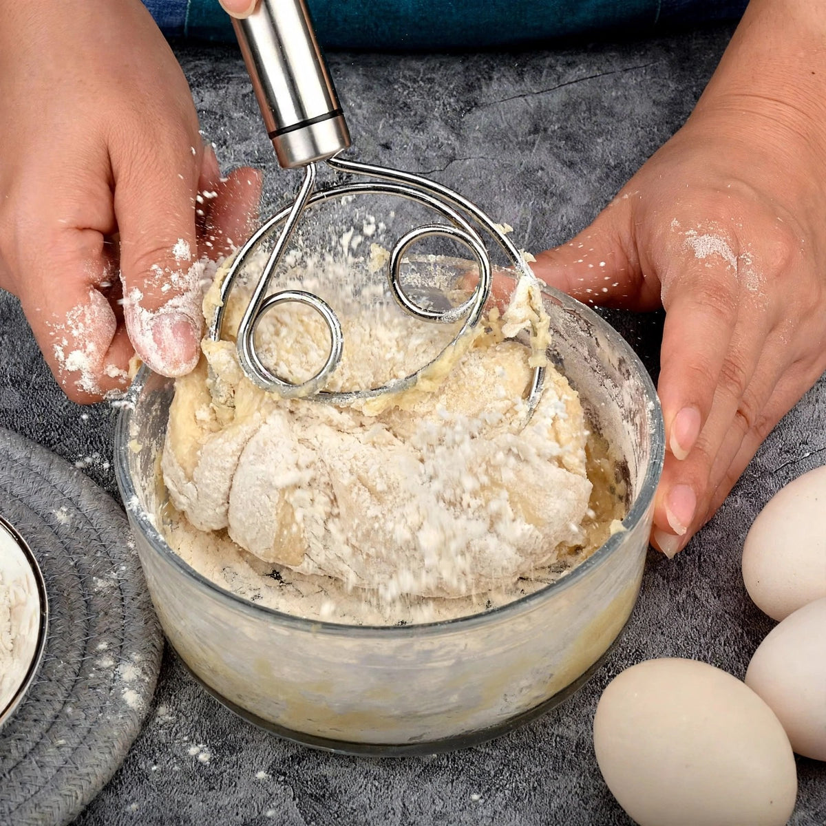 Fouet à pâte Cristal travaillant une pâte épaisse dans un bol en verre, mains farineuses