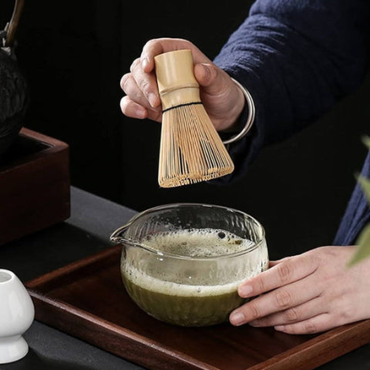 Chasen en bambou tenu au-dessus d'un bol en verre de matcha moussant, brins fins visibles
