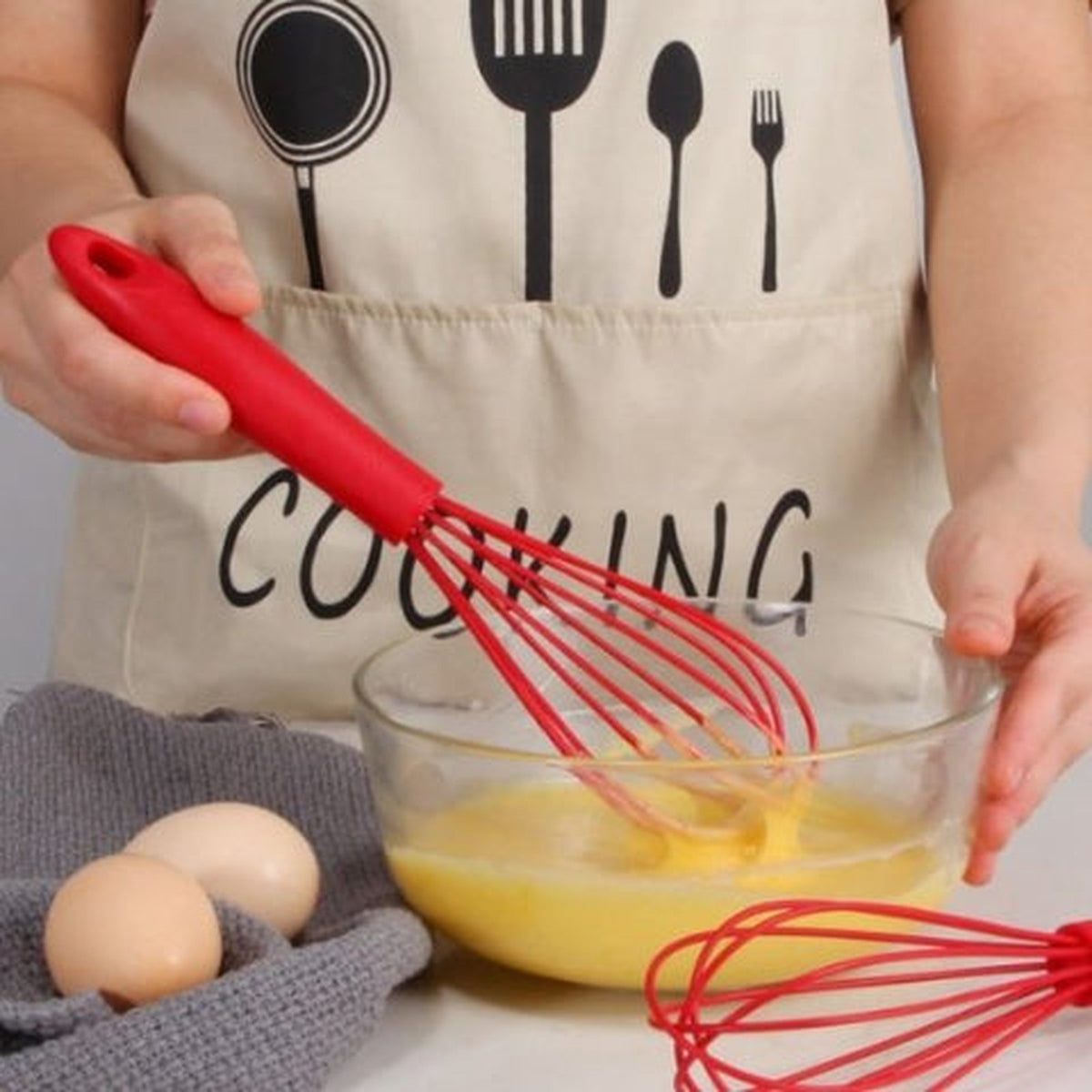 Fouet MixMaster rouge en silicone battant des œufs dans un bol en verre, tablier de cuisine